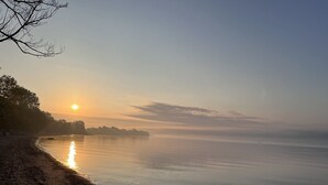 Un lac paisible au lever du soleil, bordé d’arbres, avec une étendue terrestre visible au loin.
