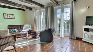 Una sala de estar con un sofá de cuero marrón, una silla de mimbre, un sillón negro, un mueble blanco con un televisor, y un ventanal con cortinas.