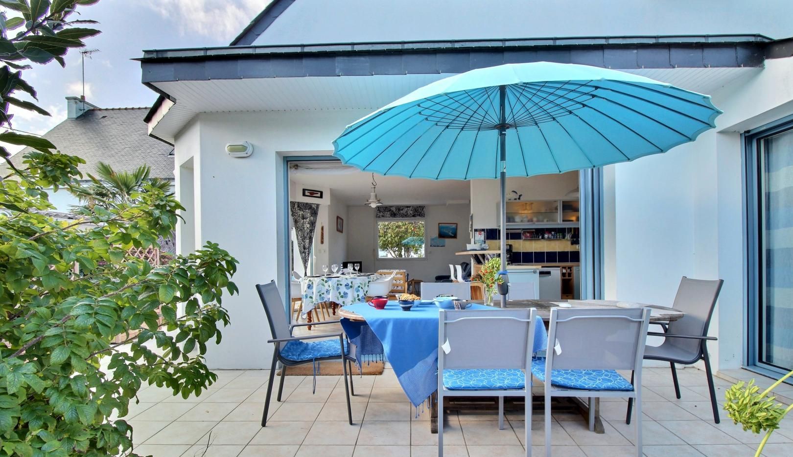 An outdoor dining area with a table set for two, a blue umbrella, and a view into the house.