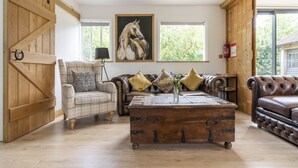 A spacious living room with a wooden floor, a large wooden coffee table, a brown leather sofa, a plaid armchair, and a large painting of a horse on the wall.