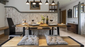 A modern dining area with a wooden table, black chairs, and grey cushions. There are hanging pendant lights and a framed picture on the wall.