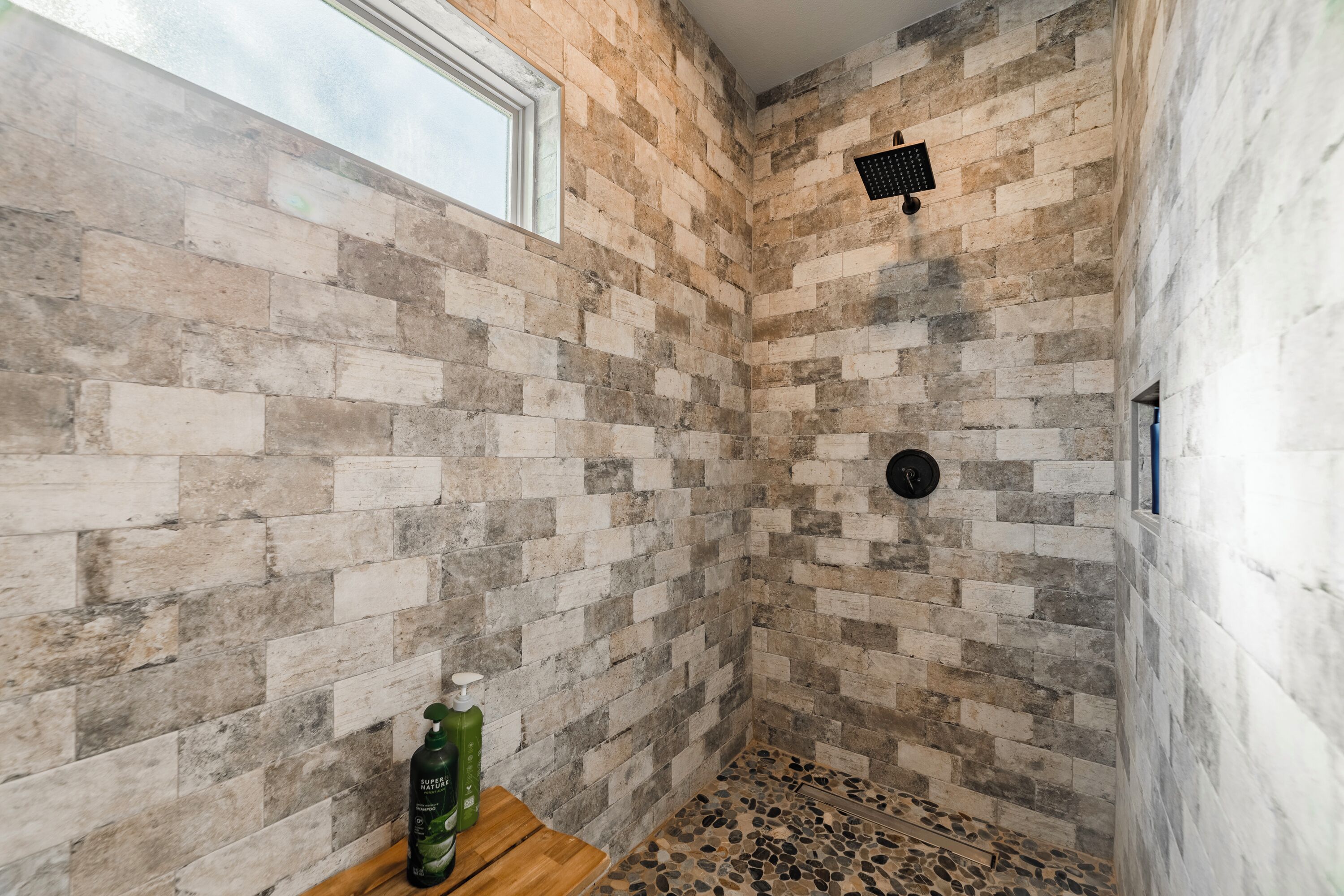 A shower with a brick wall, a window, and a wooden shelf with a bottle.