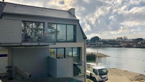 A modern two-story house with large windows and a balcony, located near a body of water.