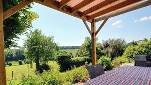 Une structure de pergola en bois, avec une table et des chaises, donnant sur un paysage verdoyant avec des arbres et un champ.