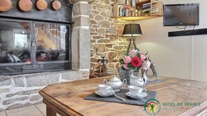 Une pièce chaleureuse avec une cheminée en pierre, une table en bois sur laquelle est disposé un service à thé, et une bibliothèque remplie de livres.