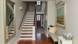 Un couloir doté de planchers en bois franc, d’un escalier, d’une console blanche sur laquelle sont posés des magazines, et d’un grand tableau encadré accroché au mur.