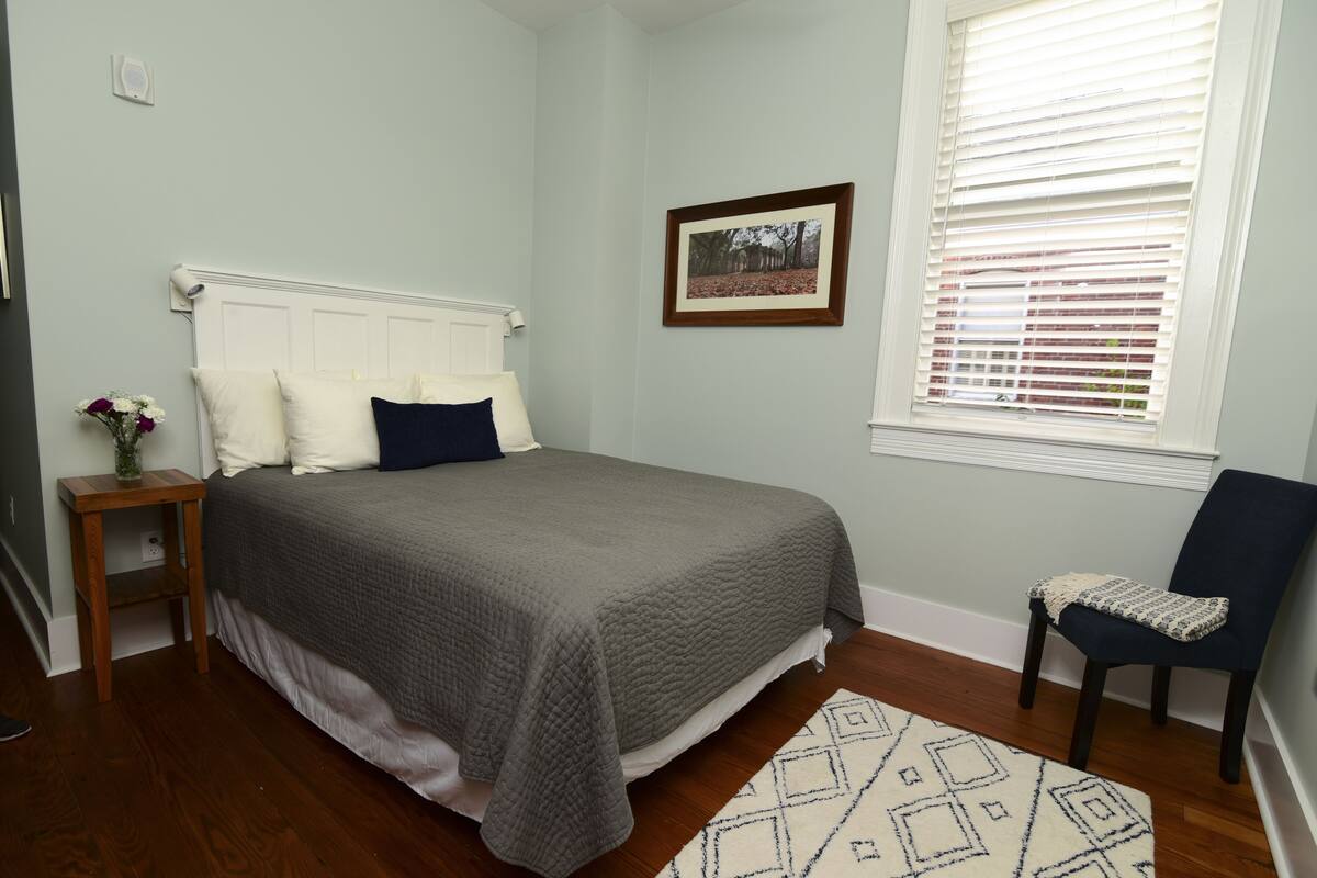Un dormitorio con cama, mesita de noche, silla, ventana con persianas e imagen enmarcada en la pared.