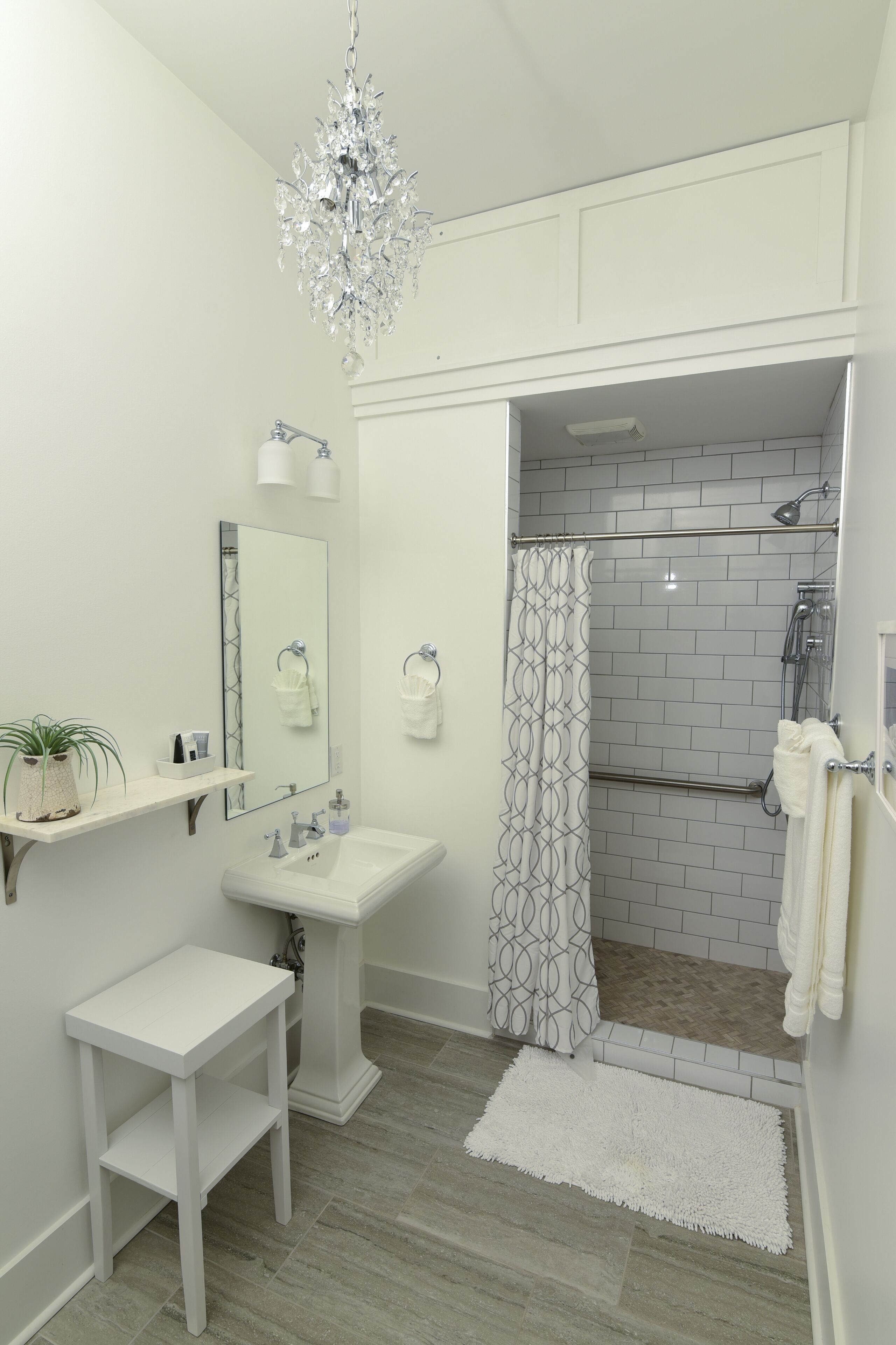 A bathroom with a pedestal sink, a mirror, a chandelier, a shower with a patterned curtain, and a white rug.