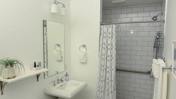 A bathroom with a pedestal sink, a mirror, a chandelier, a shower with a patterned curtain, and a white rug.