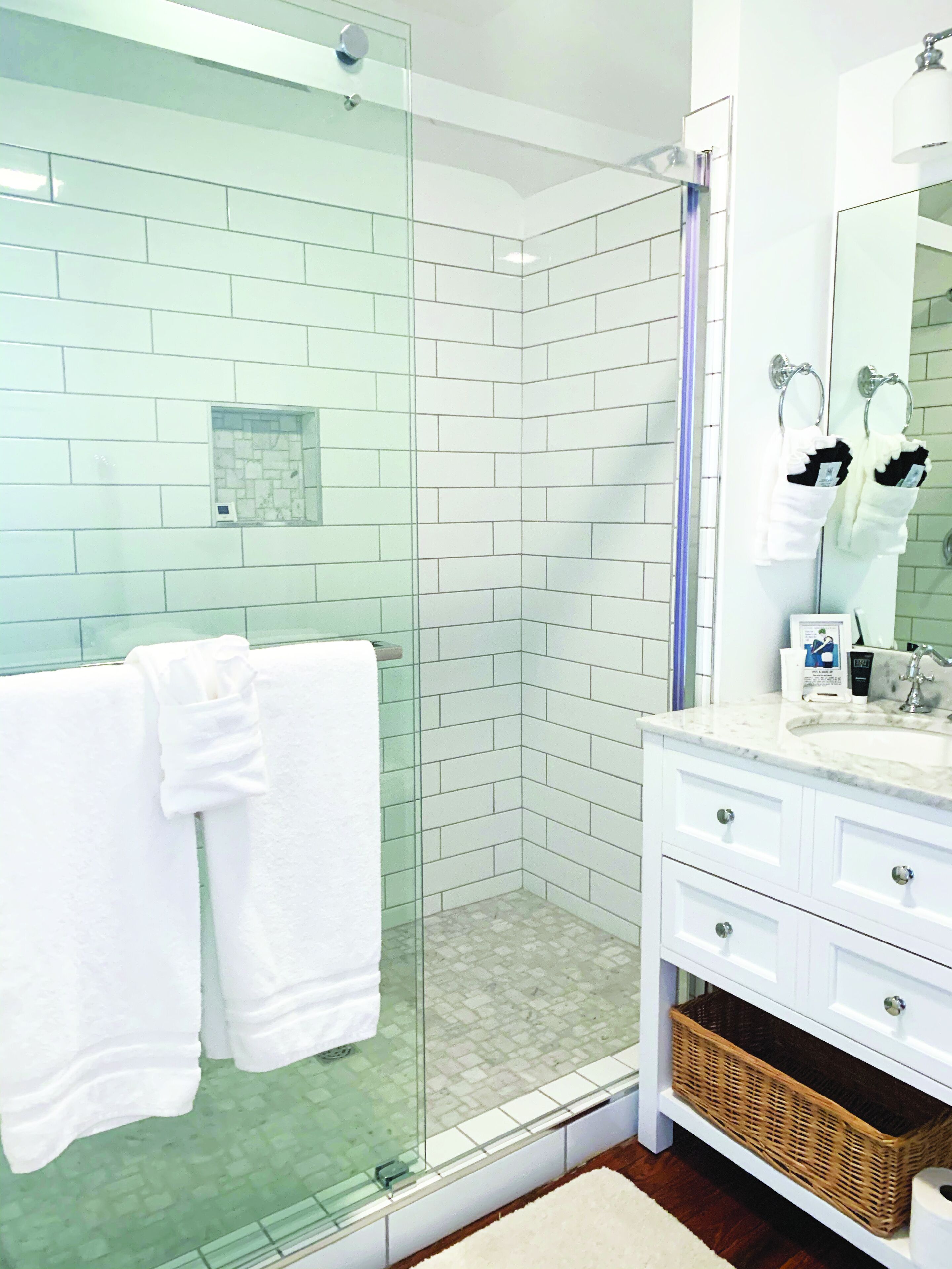 A modern bathroom with white tiled walls, a glass shower enclosure, a white vanity with drawers, and a large mirror.