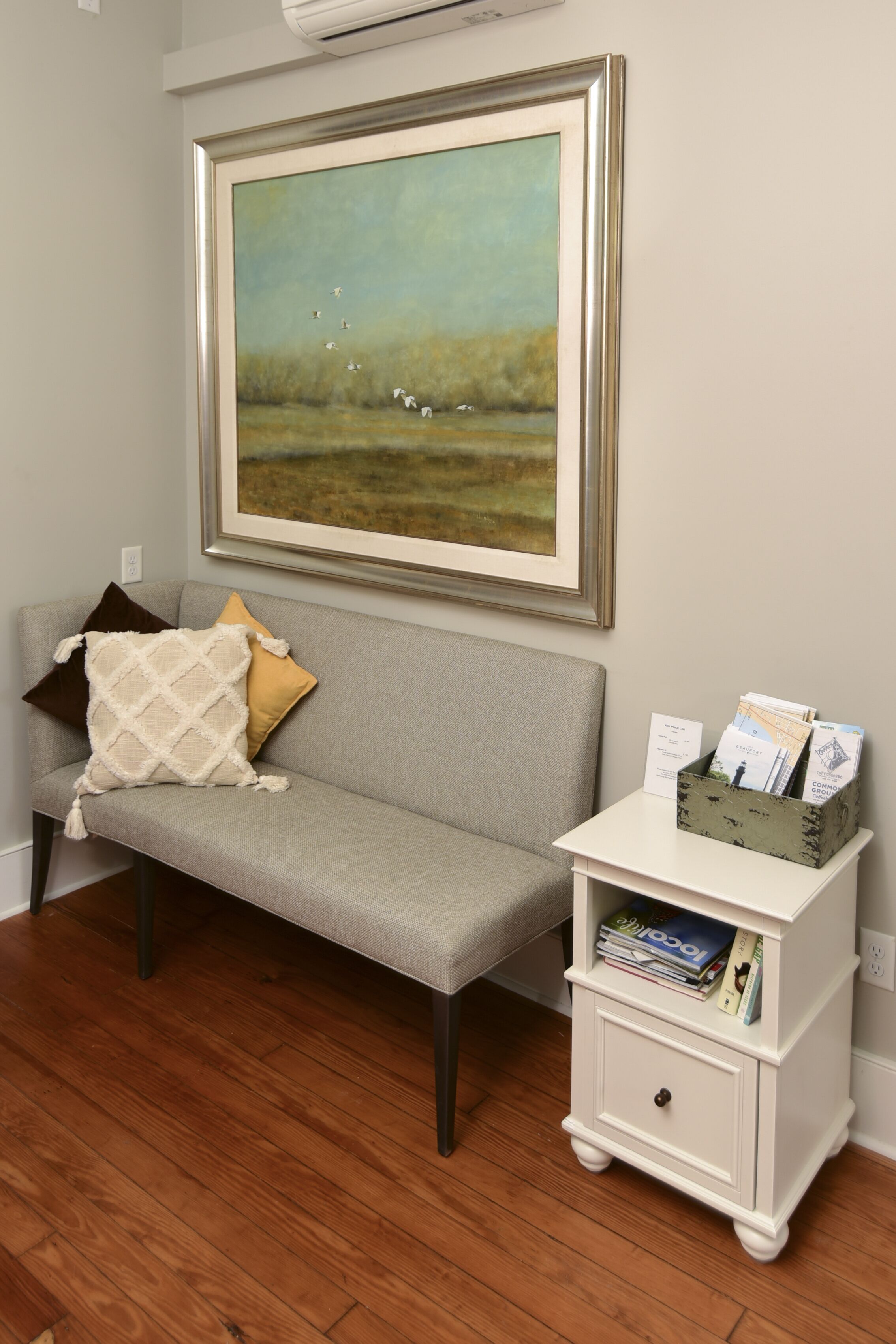 A room with a grey sofa, a framed landscape painting, a white side table with magazines, and a wooden floor.