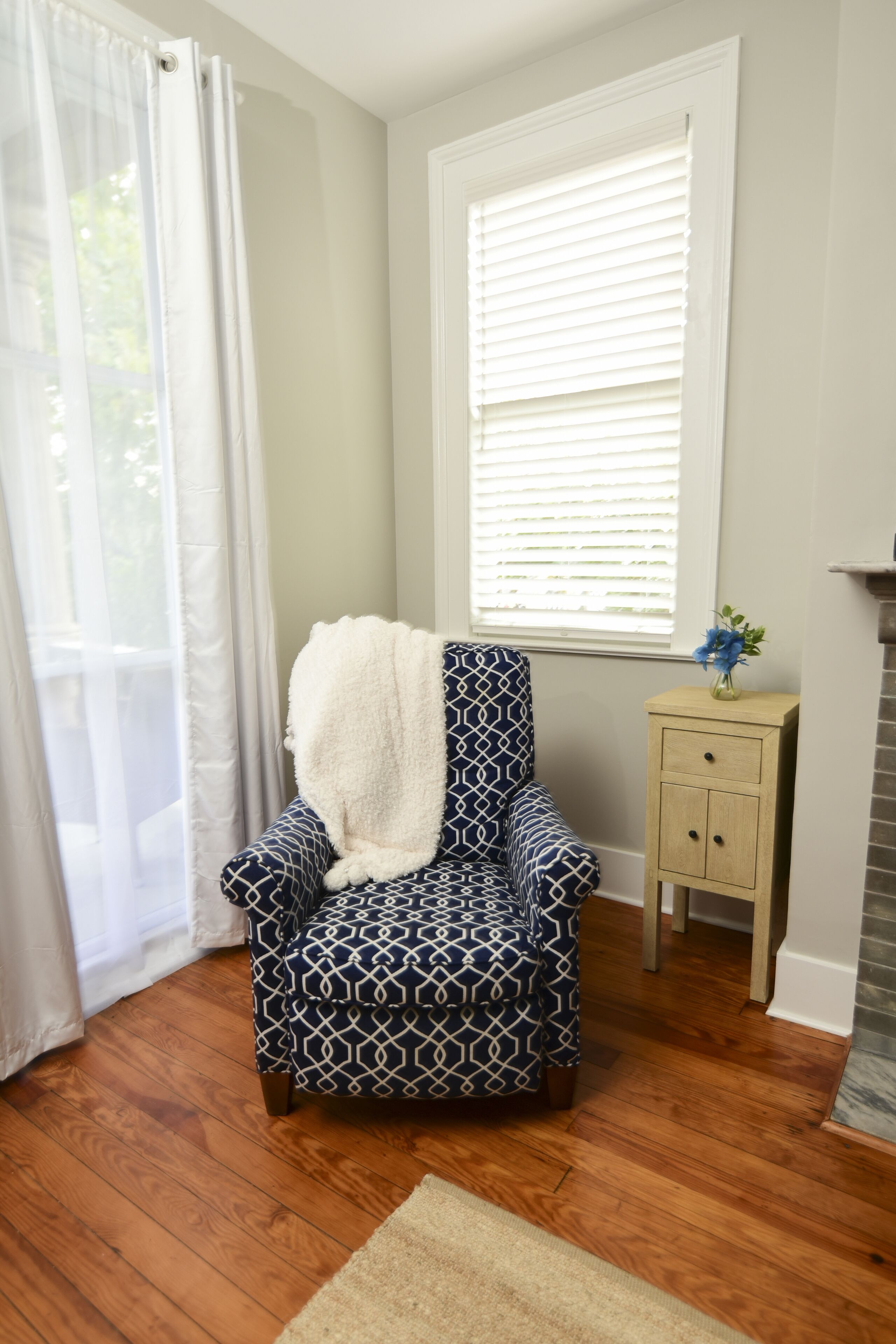 Una habitación con un sillón estampado, una pequeña mesita de madera con un florero y una ventana con persianas.