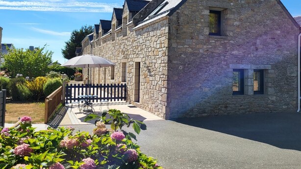 Un bâtiment en pierre avec un toit en pente, un petit porche avec une table et des chaises, et un jardin agrémenté de fleurs roses.