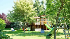 Une cabane en bois, dotée d’un toboggan et d’un mur d’escalade, entourée d’une pelouse verdoyante et luxuriante ainsi que d’arbres.