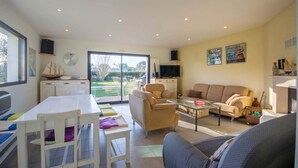A spacious living area with a sofa, a coffee table, and a television. There is a dining table with chairs and a window providing a view of the outdoors. - Les Filets de la Ria (BELZ)