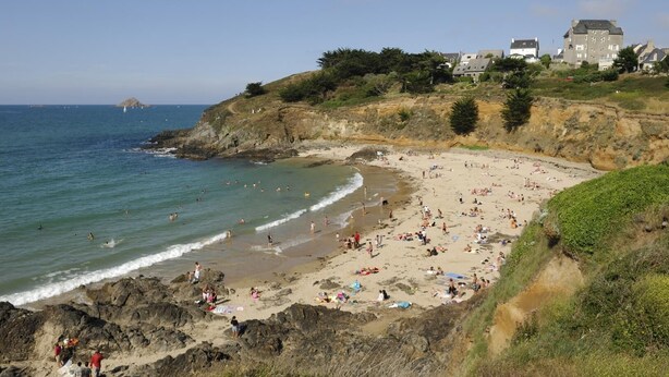 Una playa con terreno rocoso, una zona arenosa con gente y edificios al fondo.