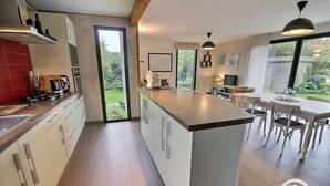 Une cuisine moderne dotée d’un îlot central, d’un coin repas avec une table et des chaises, et offrant une vue sur le jardin grâce à une porte coulissante.