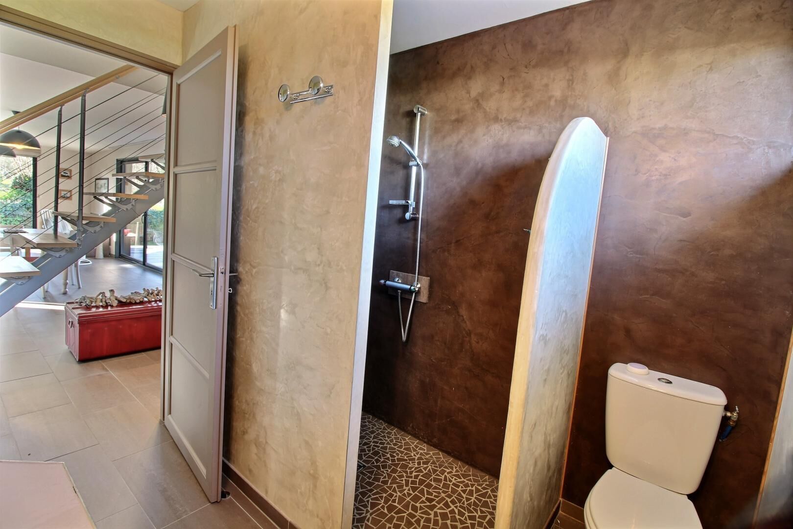 A modern bathroom with a shower, toilet, and a red bench. A staircase is visible in the background.
