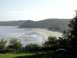 Eine Küstenlandschaft mit Sandstrand, einem grasbewachsen Vordergrund und einem bewaldeten Hügel im Hintergrund.