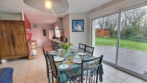 A dining area with a table set for four, a kitchen in the background, and a view of a house and garden through large sliding doors.