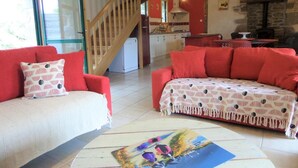 Un salon avec un canapé rouge, un canapé blanc et une table basse sur laquelle est posée une peinture. Un escalier mène à un espace cuisine doté d’une cheminée en pierre.