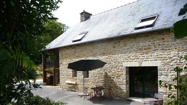 Un bâtiment en pierre doté d’un toit en pente, d’un espace extérieur abrité avec des tables et des chaises, et d’un grand parasol.