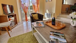 Una sala de estar moderna con un televisor de pantalla plana, un sofá cómodo, una mesa de comedor con una revista, una botella de champán y vistas al exterior a través de grandes ventanales.