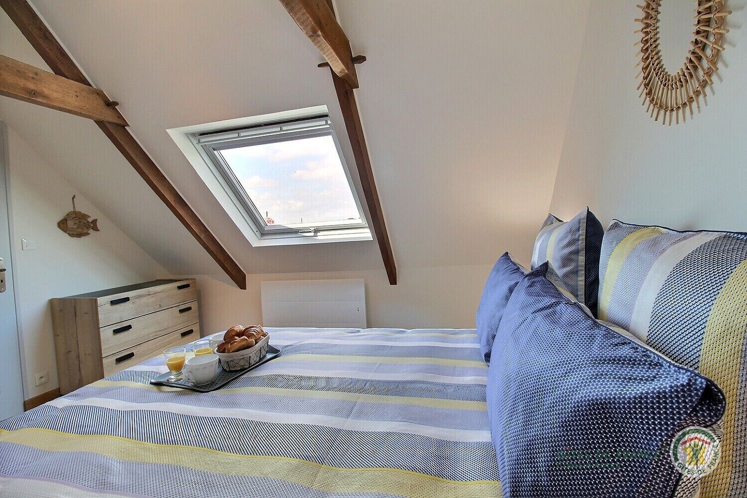 A bedroom with a sloped ceiling, a skylight, a bed with striped bedding, a bedside table with a bowl of bread and a glass of orange juice, and a wall-mounted decorative item.