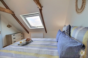 A bedroom with a sloped ceiling, a skylight, a bed with striped bedding, a bedside table with a bowl of bread and a glass of orange juice, and a wall-mounted decorative item. - Ty Coz - YVIAS - Gîte (YVIAS)