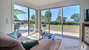 A modern living room with a sofa, a coffee table, and a TV. Large sliding glass doors lead to an outdoor area with a barbecue and a view of the sea.