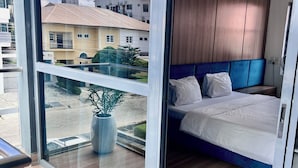 A modern bedroom with a large window, a bed, a bedside table, and a potted plant. - ATLANTA @ AMERICA LUXURY EXTENDED STAY (Lagos)