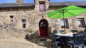 Un bâtiment en pierre avec une porte rouge, un petit balcon et une table entourée de chaises, surmontée d’un parasol vert.