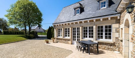 Un bâtiment en pierre avec une terrasse carrelée, une table entourée de chaises et un arbre en arrière-plan.