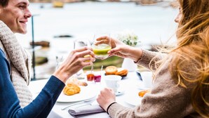 Una pareja brindando con bebidas en una mesa con pasteles, con vista a una masa de agua.