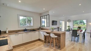 A modern kitchen with a wooden countertop, white cabinets, and a bar area with stools. There is a dining area with a table and chairs, and a view of greenery outside. - du Talbert - Pleubian (PLEUBIAN)