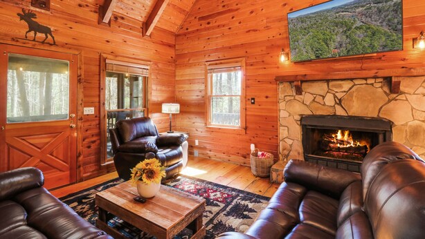 A cozy log cabin interior with a fireplace, leather sofas, and a large TV mounted on the wall. - Escape to Moose Crossing – A Cozy Cabin Retreat in Blue Ridge, GA! (Blue Ridge)