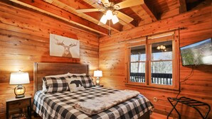 A wooden cabin bedroom with a bed, two bedside lamps, a ceiling fan, a mounted TV, and a window with a view of the outdoors. - Escape to Moose Crossing – A Cozy Cabin Retreat in Blue Ridge, GA! (Blue Ridge)
