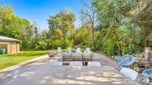 Une terrasse extérieure avec une grande table et des chaises, entourée d’arbres et d’un mur en pierre.