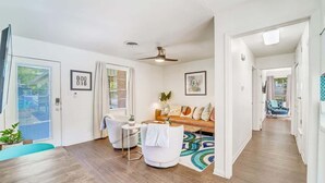 Un salon moderne avec un canapé, des fauteuils et une table basse. On y trouve une porte avec une pancarte, un ventilateur de plafond et une fenêtre.