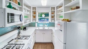 Une cuisine de taille réduite, avec des meubles blancs, une cuisinière à gaz, un évier et un réfrigérateur. Des étagères sont remplies de divers ustensiles de cuisine.