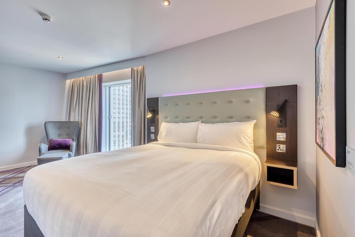 Une chambre d’hôtel moderne avec un grand lit, une chaise, une étagère fixée au mur et une fenêtre avec des rideaux.