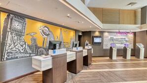 Een hotel receptie met een groot muurdecoratie van Londense bezienswaardigheden, een digitaal informatiepaneel en een zelfbedieningskiosk.