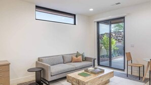 Sala de estar moderna com sofá cinza, mesa de centro de madeira e uma pequena mesa lateral. Há uma porta de vidro deslizante que dá para uma área externa com plantas.