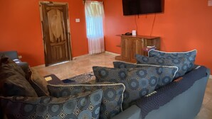 Un salon aux murs orange, doté d’un ventilateur de plafond, d’un téléviseur fixé au mur, d’un meuble en bois et de coussins à motifs sur un canapé.