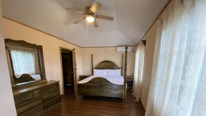 Une chambre avec un plancher en bois, un lit avec une tête de lit, une commode, un ventilateur de plafond et une fenêtre avec des rideaux légers.