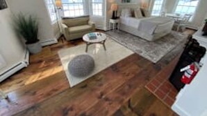 A spacious living room with a large sofa, a coffee table, and a rug. There are two windows providing natural light, and a fireplace is visible in the background.