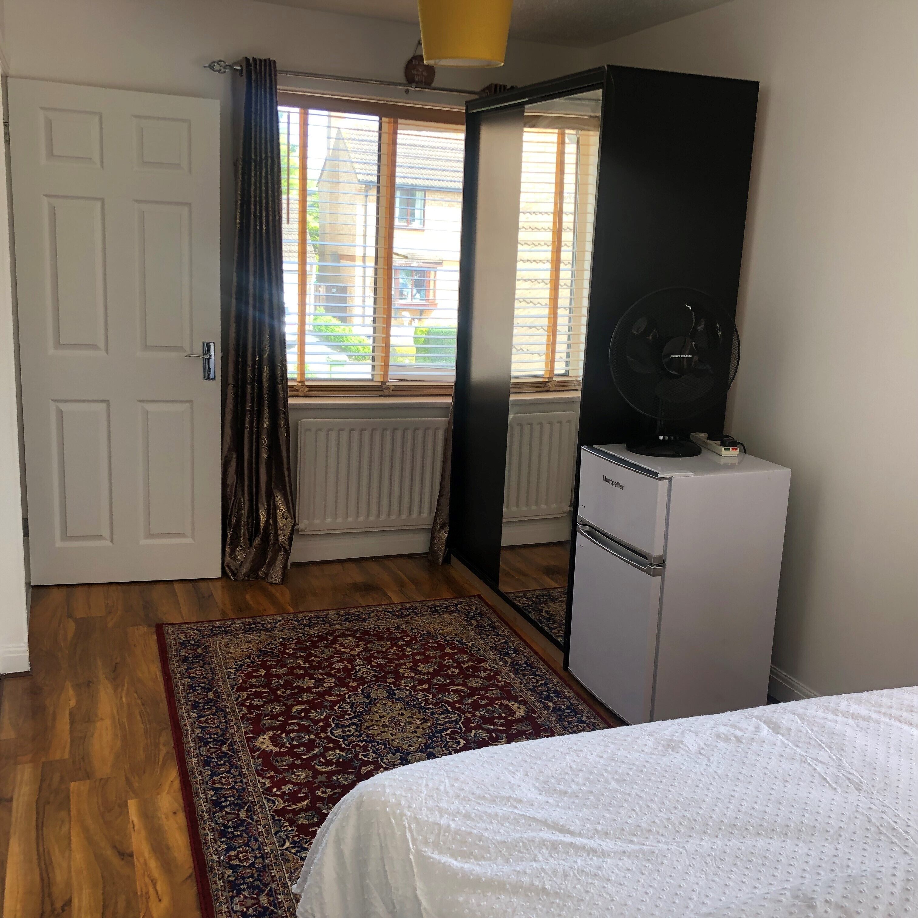 A bedroom with a bed, a radiator, a window with blinds, a fan, and a door.