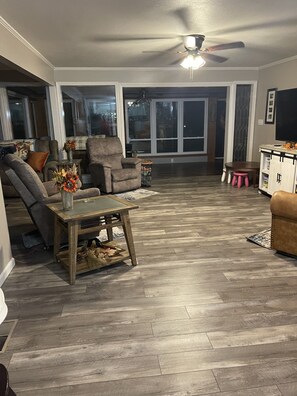 A spacious living room with a large window, a ceiling fan, and a wooden floor.