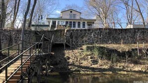 Un puente de piedra con barandilla metálica que lleva a una casa de fachada blanca y balcón.