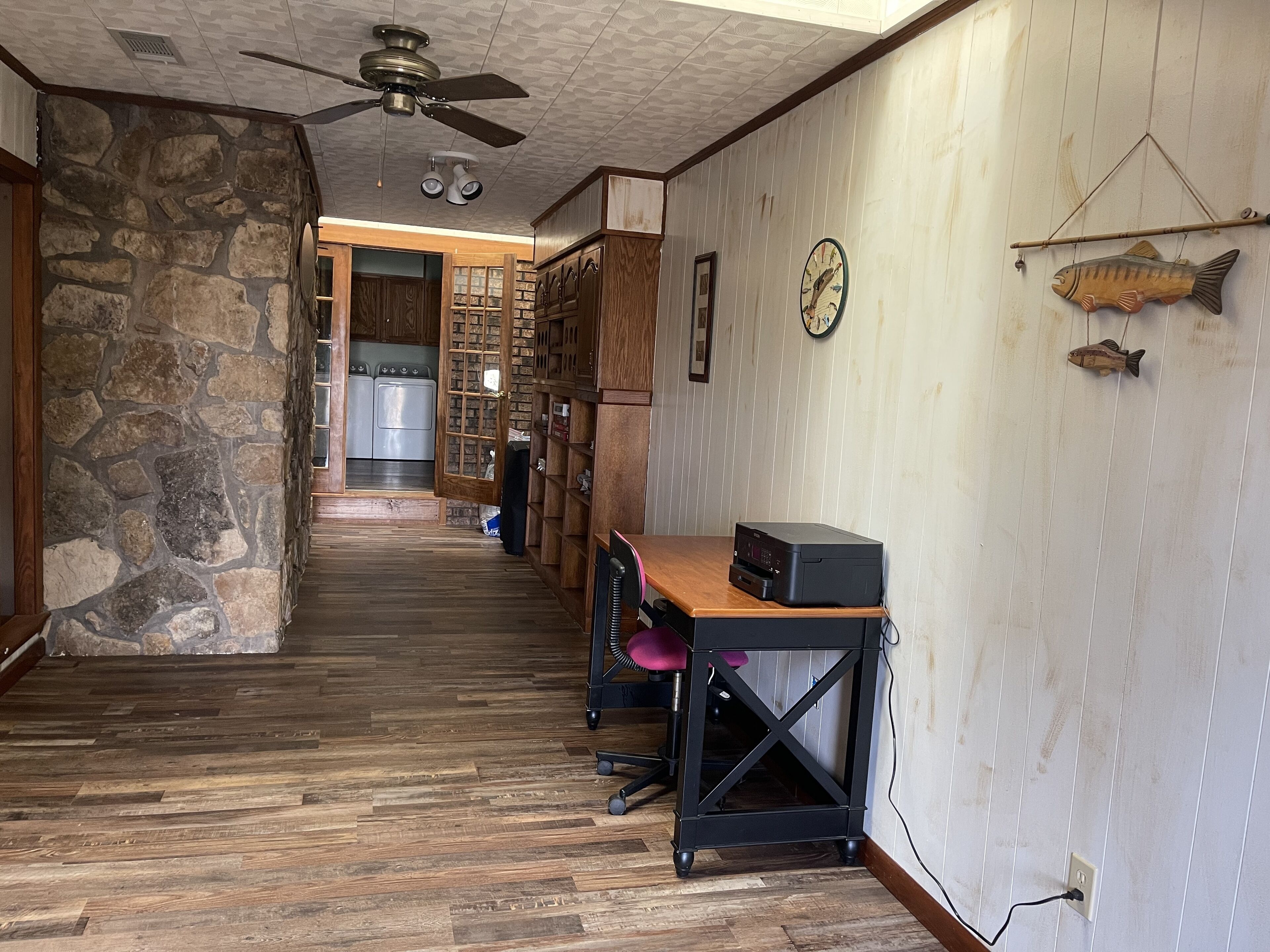 Un pasillo con pared de piedra, piso de madera, ventilador de techo, un reloj y una decoración de pez.
